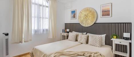 Un lit double avec une couverture beige, une table de chevet avec une lampe, un radiateur fixé au mur et une fenêtre avec des rideaux.