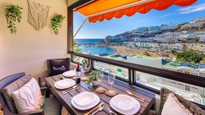 Un balcón con mesa lista para comer, vista a un pueblo costero y un tapiz de macramé en la pared.