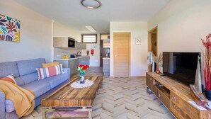 Una sala moderna con sofá, mesa de centro, televisor y una cocina al fondo.