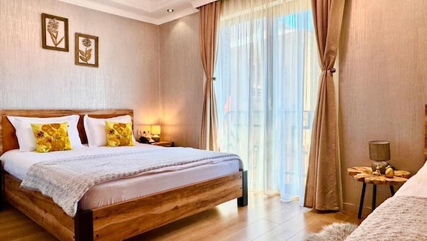 A bedroom with a wooden bed, two bedside tables, a nightstand, a window with curtains, and framed wall art. - Gold Tower in Mestia (Mestia)