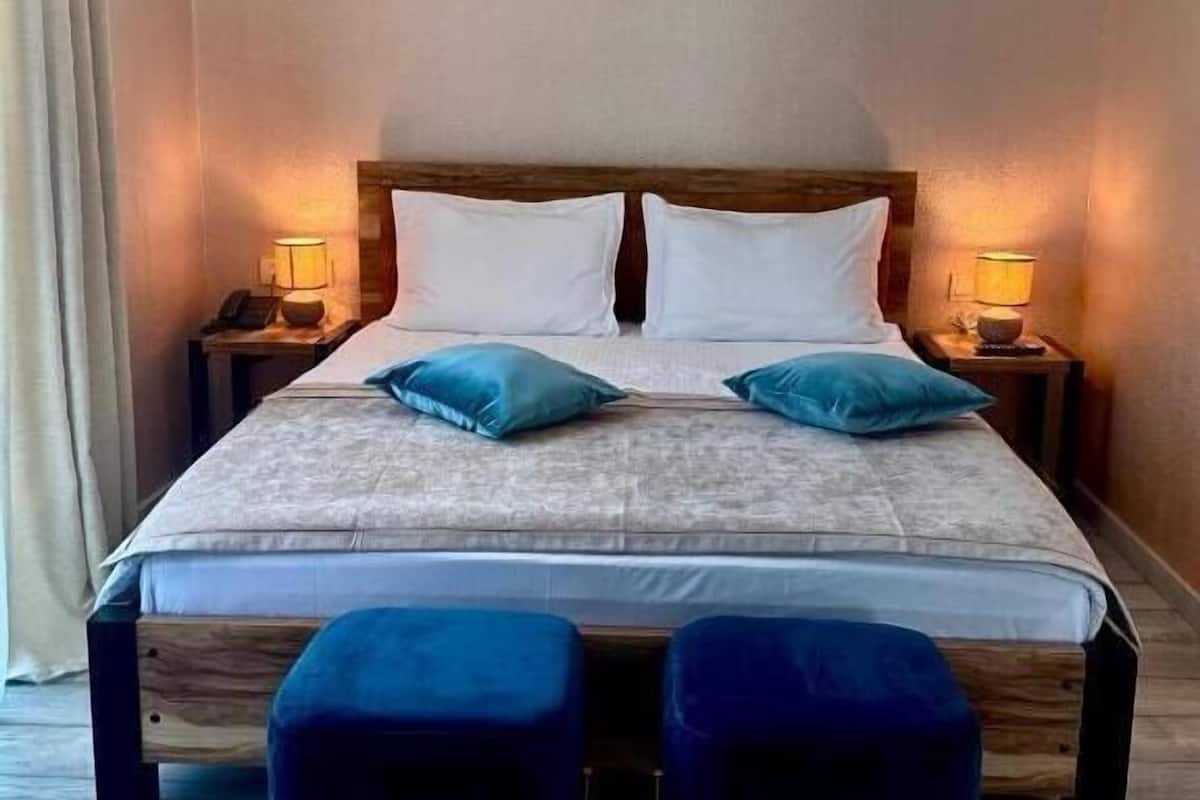 A hotel room with a bed, two bedside tables with lamps, and a pair of blue ottomans.