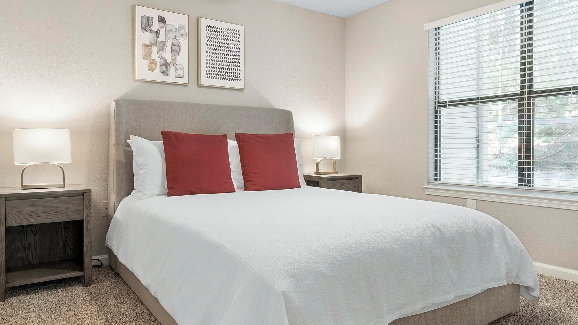 A neatly made bed with white bedding and two red pillows, flanked by nightstands with lamps, in a room with a large window and two wall-mounted artworks.