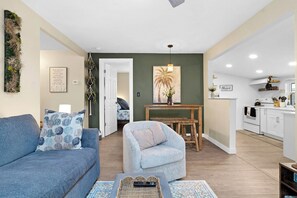 Un salon comprenant un canapé, un fauteuil et une table basse. Il y a également un coin cuisine avec des meubles blancs et une table à manger.