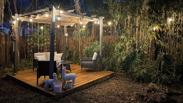 Una terrazza in legno con un'area salotto coperta, circondata da fitta vegetazione e luci decorative.
