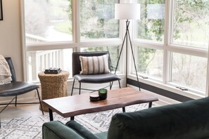 A room with a large window, a floor lamp, a wooden table, a green sofa, and a black chair with a striped pillow. - 7 Acres Stunning Custom "Treehouse" on the New River - 2 King Beds, 2 twin lofts (Boone)