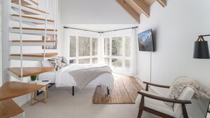 Ein Schlafzimmer mit einer Holztreppe, einem Bett, einem Schreibtisch, einem Stuhl und einem großen Fenster mit Blick auf Bäume.