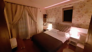 Quarto de hotel com cama, cortinas, mesa de cabeceira, abajur e janela.