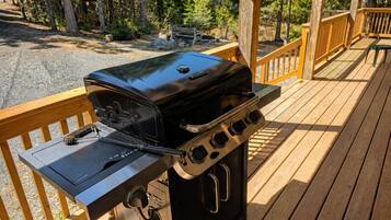 Una parrilla de barbacoa en una terraza de madera con vistas a árboles y un sendero de grava.