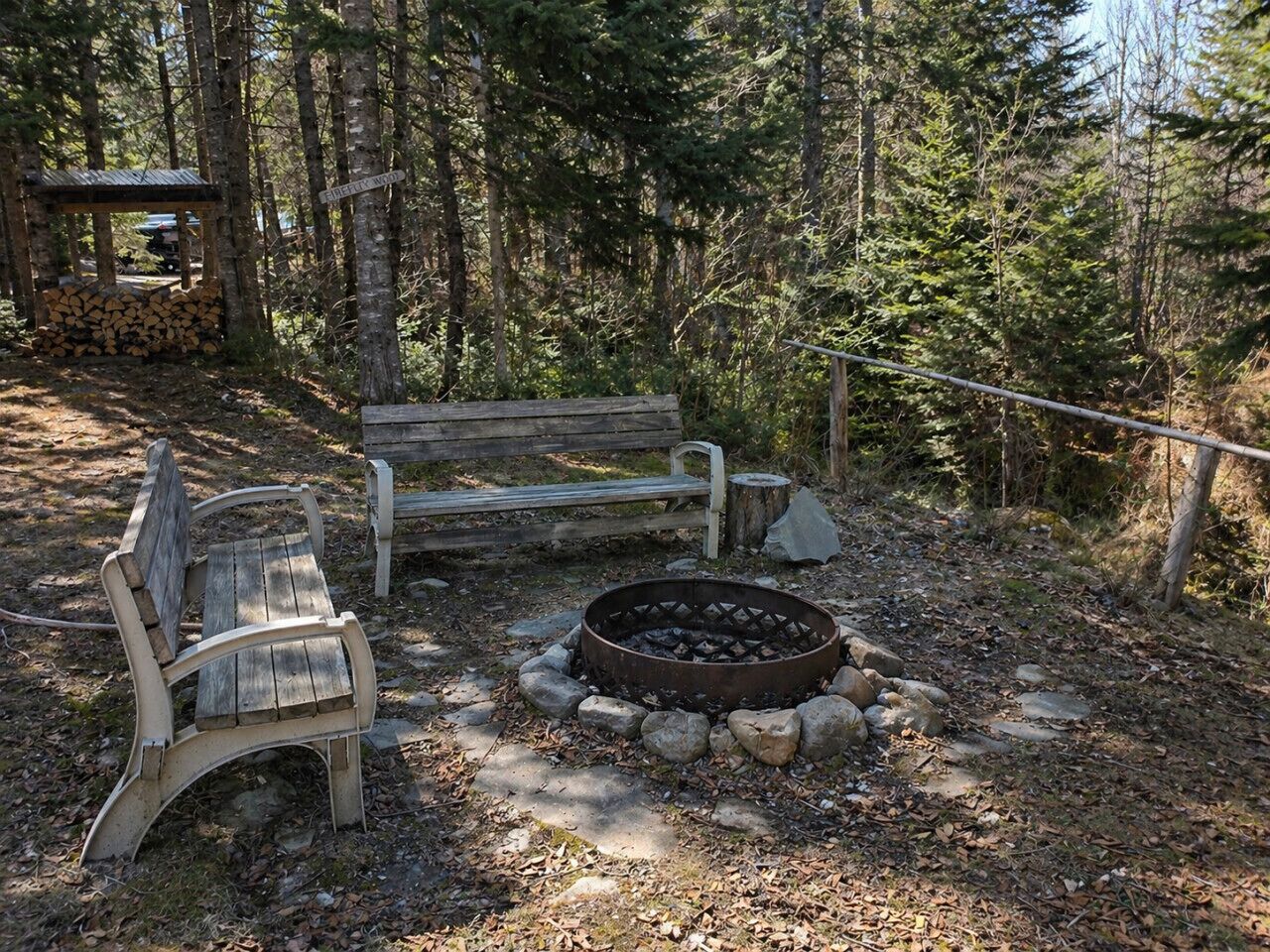 A forested area with a fire pit, two benches, and a wooden structure.