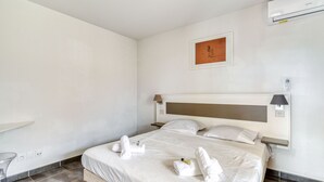 A hotel room with a bed, a wall-mounted air conditioner, a framed picture, and a bedside table.