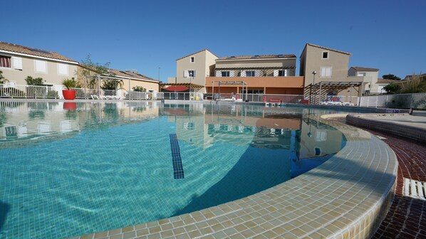 A swimming pool with tiled edges, surrounded by a white fence, and buildings in the background.