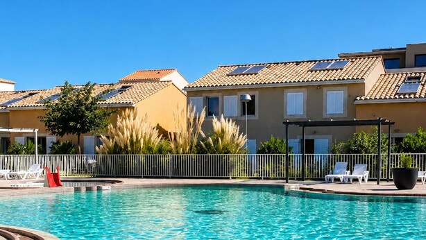 A swimming pool with a tiled edge, surrounded by a white fence, and adjacent to buildings with a tiled roof.