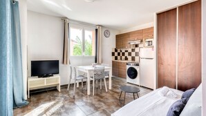A compact room with a bed, a TV, a dining table with chairs, a washing machine, and a kitchenette.