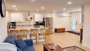 A modern kitchen with white cabinets, a central island with bar stools, a dining area with a sofa and coffee table, and a hallway with a door.