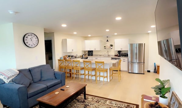 A modern living room with a sofa, coffee table, and a dining area with wooden chairs and a kitchen with white cabinets and stainless steel appliances.
