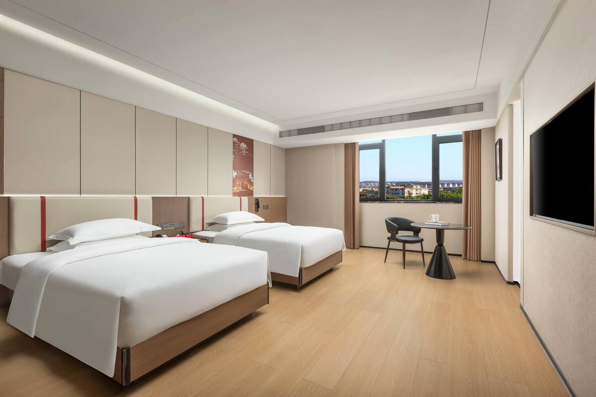Habitación de hotel con dos camas, un escritorio y un amplio ventanal con vistas a la ciudad.
