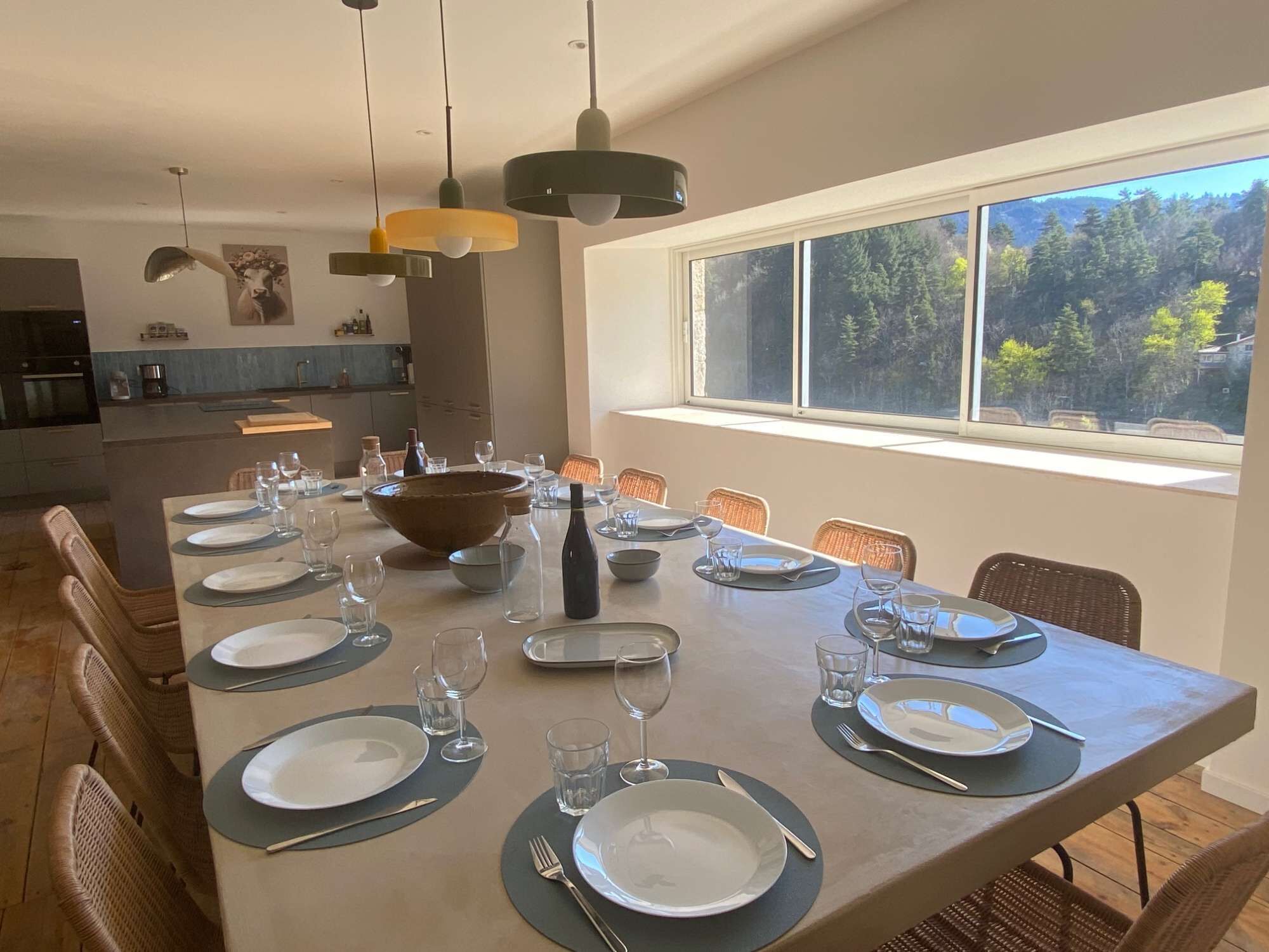Un espace repas avec une longue table dressée pour un repas, entourée de chaises en osier, et une grande fenêtre offrant une vue sur un paysage boisé.
