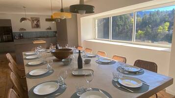 Un espace repas avec une longue table dressée pour un repas, entourée de chaises en osier, et une grande fenêtre offrant une vue sur un paysage boisé.