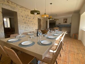 A long dining table set with plates, glasses, and cutlery, flanked by wicker chairs, in a room with stone walls and a kitchen in the background. - Les murmures de l'Eyrieux, a holiday cottage for 15 people with an indoor swimming pool (Saint Martin de Valamas)