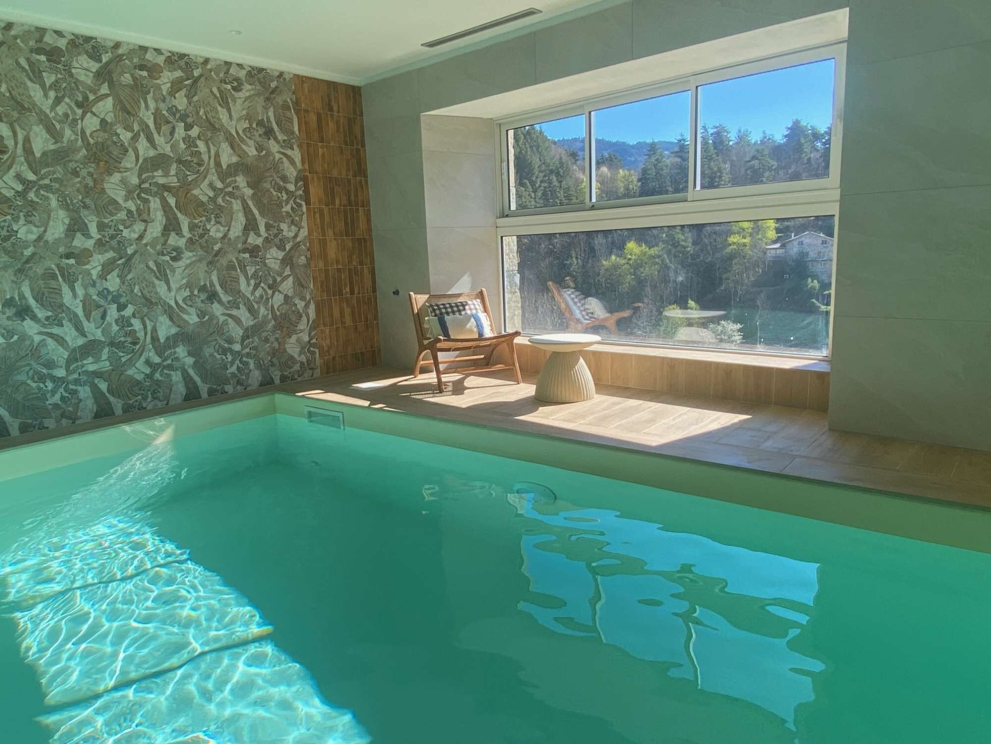 Une piscine à l’eau claire, une chaise en bois et une vue sur une forêt, visible à travers une fenêtre.