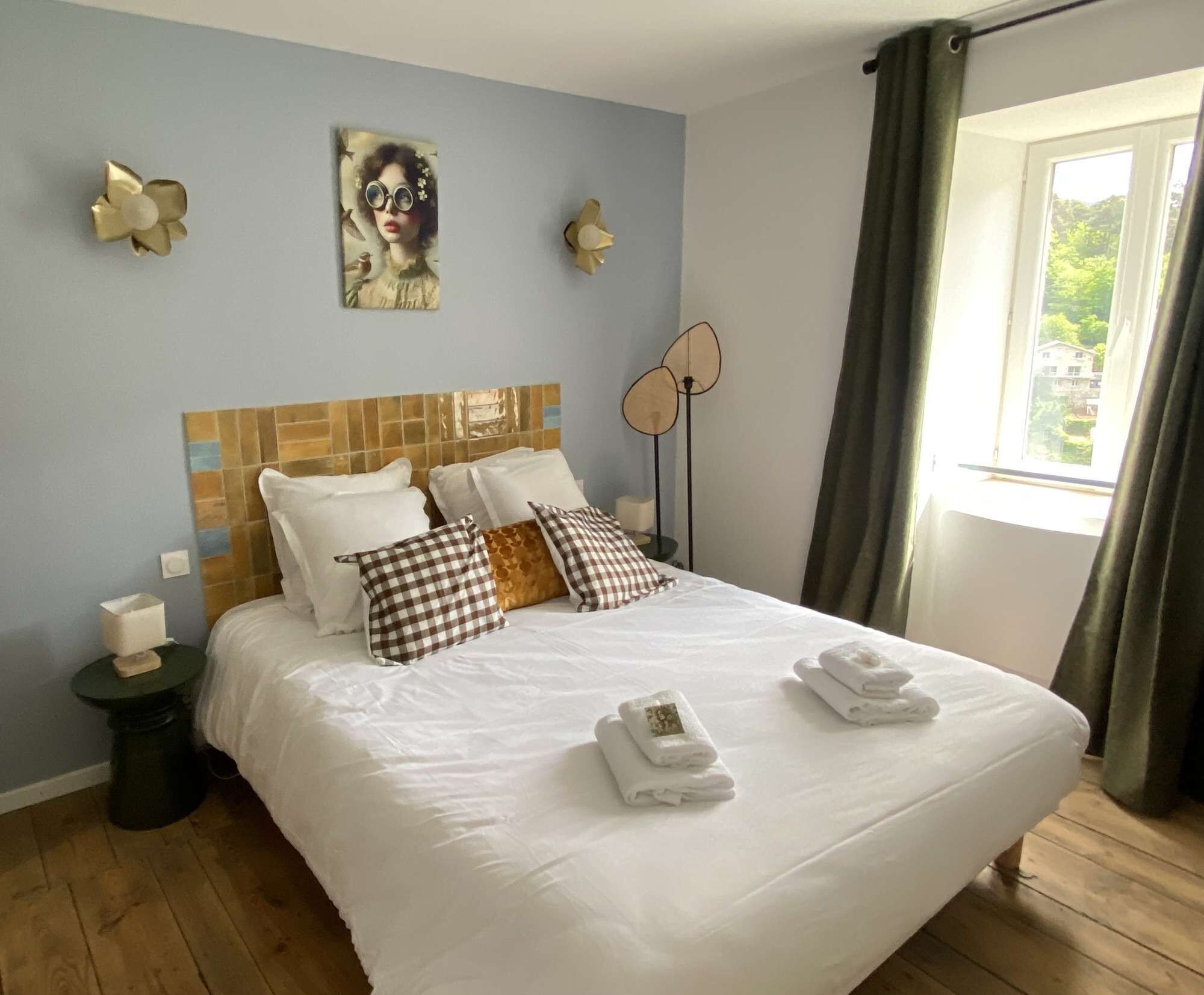 Une chambre d’hôtel avec un grand lit, une tête de lit ornée de motifs en carreaux, deux appliques murales, une fenêtre avec des rideaux et une petite table de chevet sur laquelle se trouve une boîte de mouchoirs.
