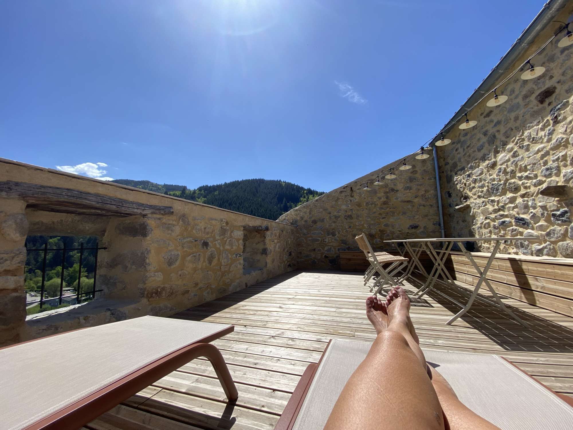 A sunny outdoor patio with stone walls, a wooden deck, and a table and chair.