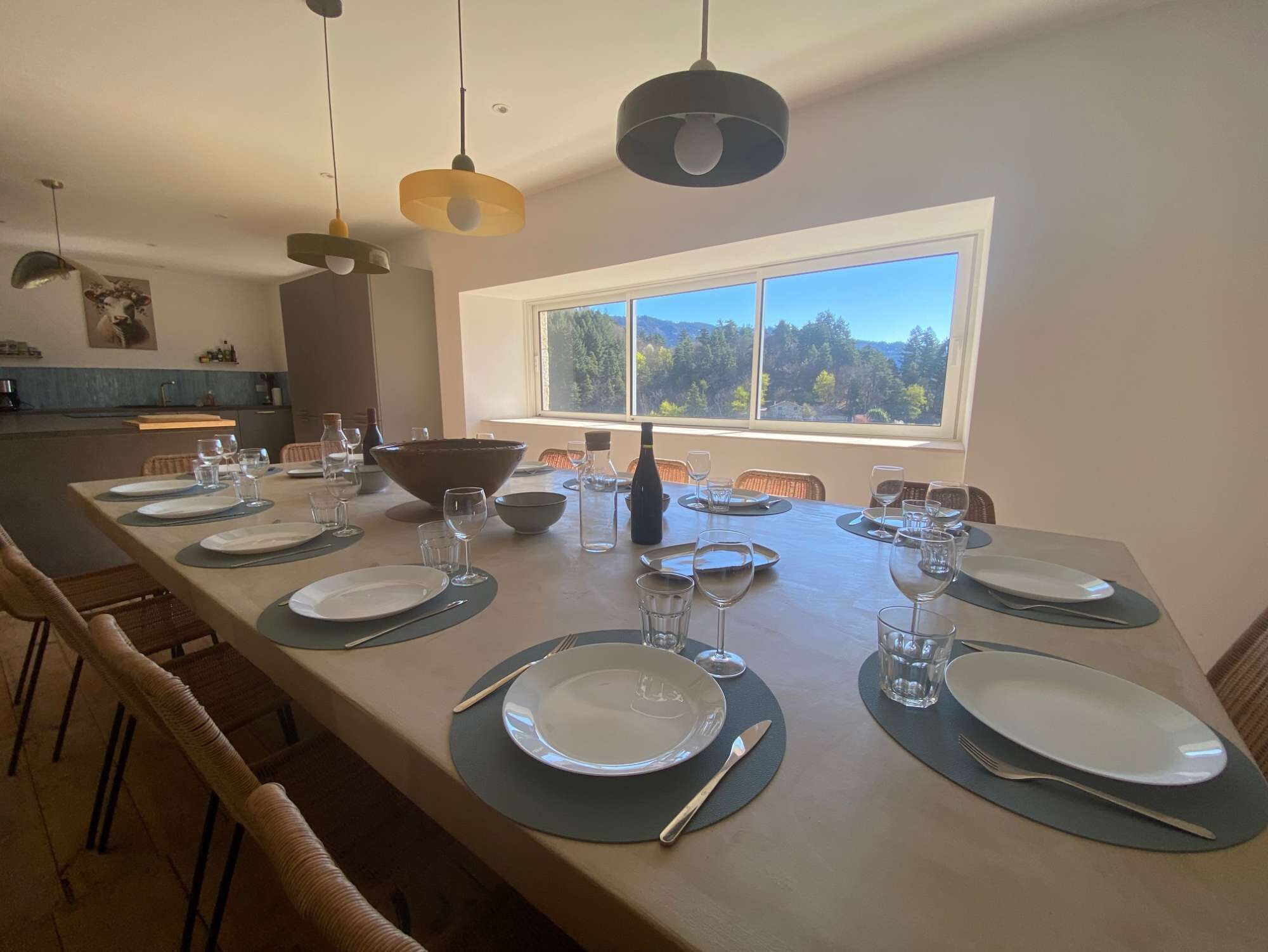 Comedor con una larga mesa dispuesta para una comida, una ventana con vista a un bosque y lámparas colgantes arriba.