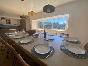 Uma sala de jantar com uma mesa comprida posta para refeição, uma janela com vista para a floresta e luminárias pendentes.