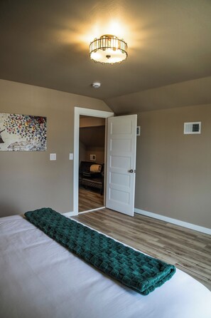 Een slaapkamer met een bed, een groene deken, een plafondlamp, een deur en een muur met een schilderij.