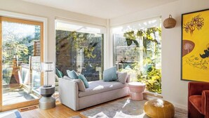 A modern living room with a sofa, a small table, and a chair. There are large windows with a view of greenery outside. - Woodstock Waterfall Hideaway · Walk To Tinker St! (Woodstock)