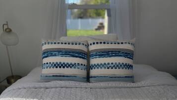 A bed with a white quilt and two patterned pillows, a bedside lamp, and a window with a view of a yard.