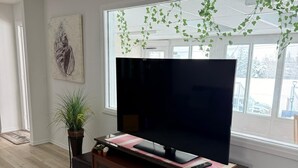 A living room with a large flat-screen TV on a wooden entertainment unit, a potted plant, a framed artwork, and a view of the outdoors through a window. - Convenient Location Near Large Shopping Mall and Train Station. (Calgary)