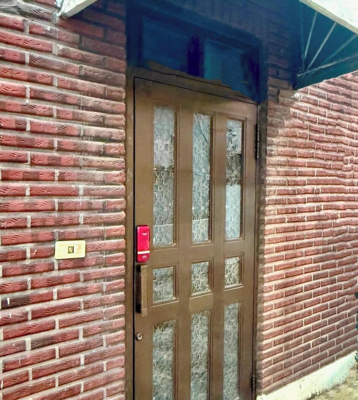 A brown door with a glass panel, a red handle, and a light switch on the brick wall to the left.