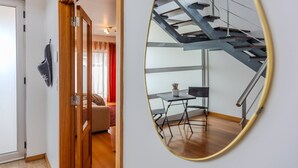 Un interior moderno con una puerta de madera, un espejo que refleja una escalera y un área de comedor con una mesa y sillas.