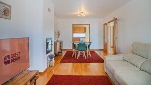 Una sala de estar moderna con un sofá, un comedor con mesa y sillas, un televisor y una cocina visible a través de una puerta abierta.