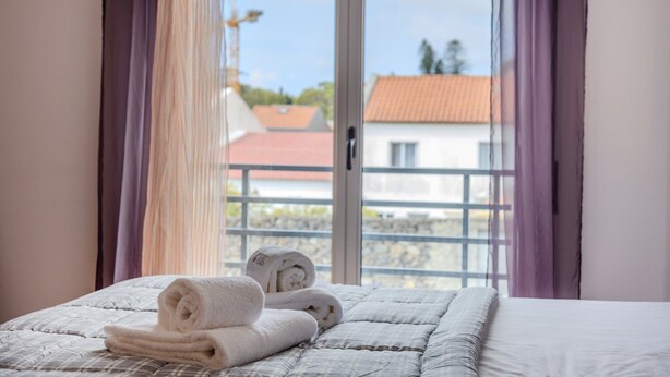 Una cama con toallas dobladas, una ventana con vistas a edificios y cortinas moradas.