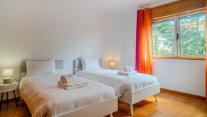 Habitación de hotel con dos camas, una ventana con cortinas, una mesita de noche con lámpara y vista a zonas verdes.