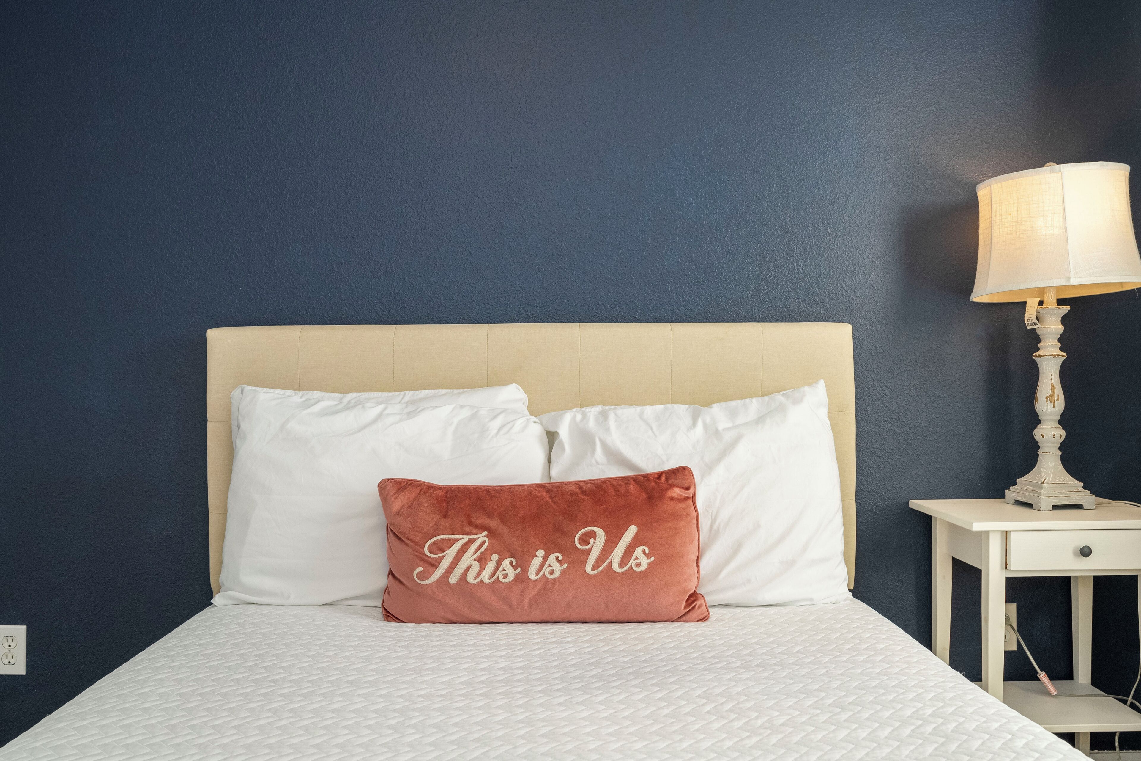 A bed with a white headboard, a single rose gold pillow with the text "This is Us," and a bedside table with a lamp.