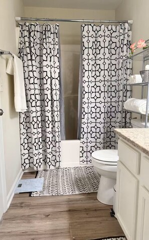 A bathroom with a shower curtain, a toilet, a sink, and a patterned rug. - Cozy Corner Cottage (Raleigh)