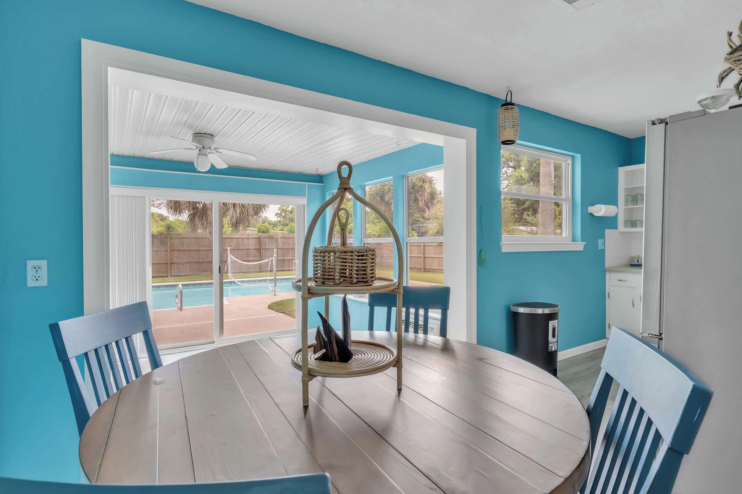 A room with a round table, chairs, a ceiling fan, a wicker chair, a trash can, and a view of a backyard with a pool.