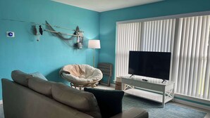 En stue med en beige sofa, en hvid lænestol, et fjernsyn på et hvidt skab, en lampe og et vindue med persienner.