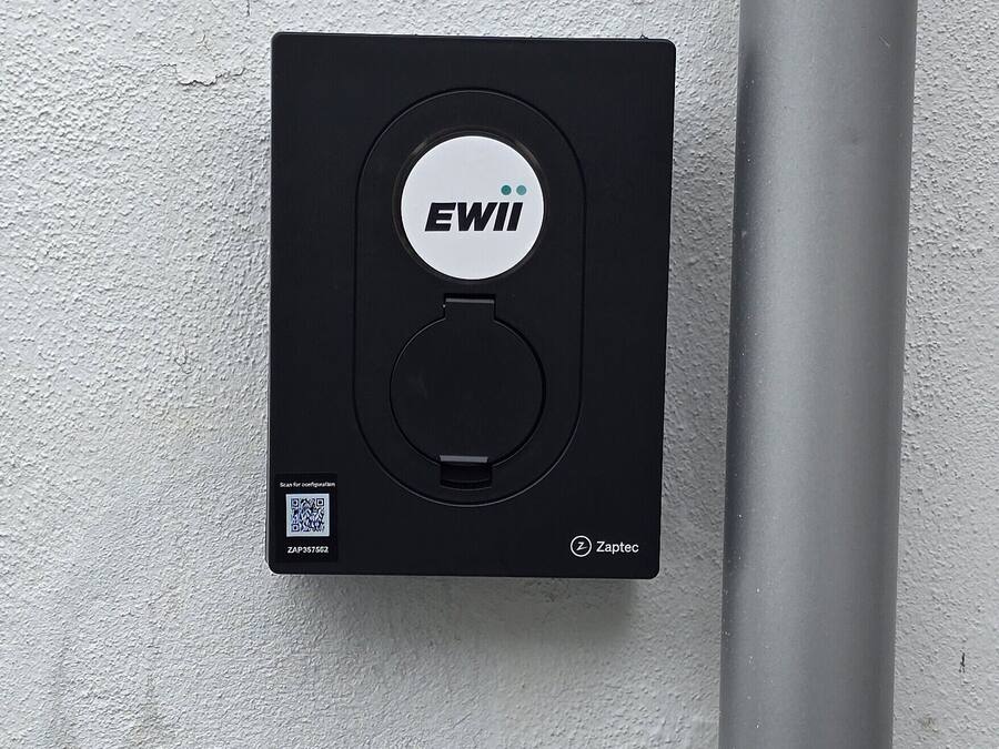 A black device with the 'EWII' logo, a QR code, and the 'Zaptec' brand on a textured wall.