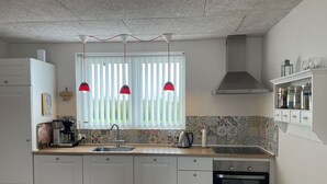 Una cocina con armarios blancos, un lavamanos de acero inoxidable, horno empotrado y ventana con persianas verticales.