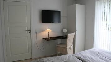 A bedroom with a bed, a desk with a lamp, a chair, a television on the wall, and a wardrobe.