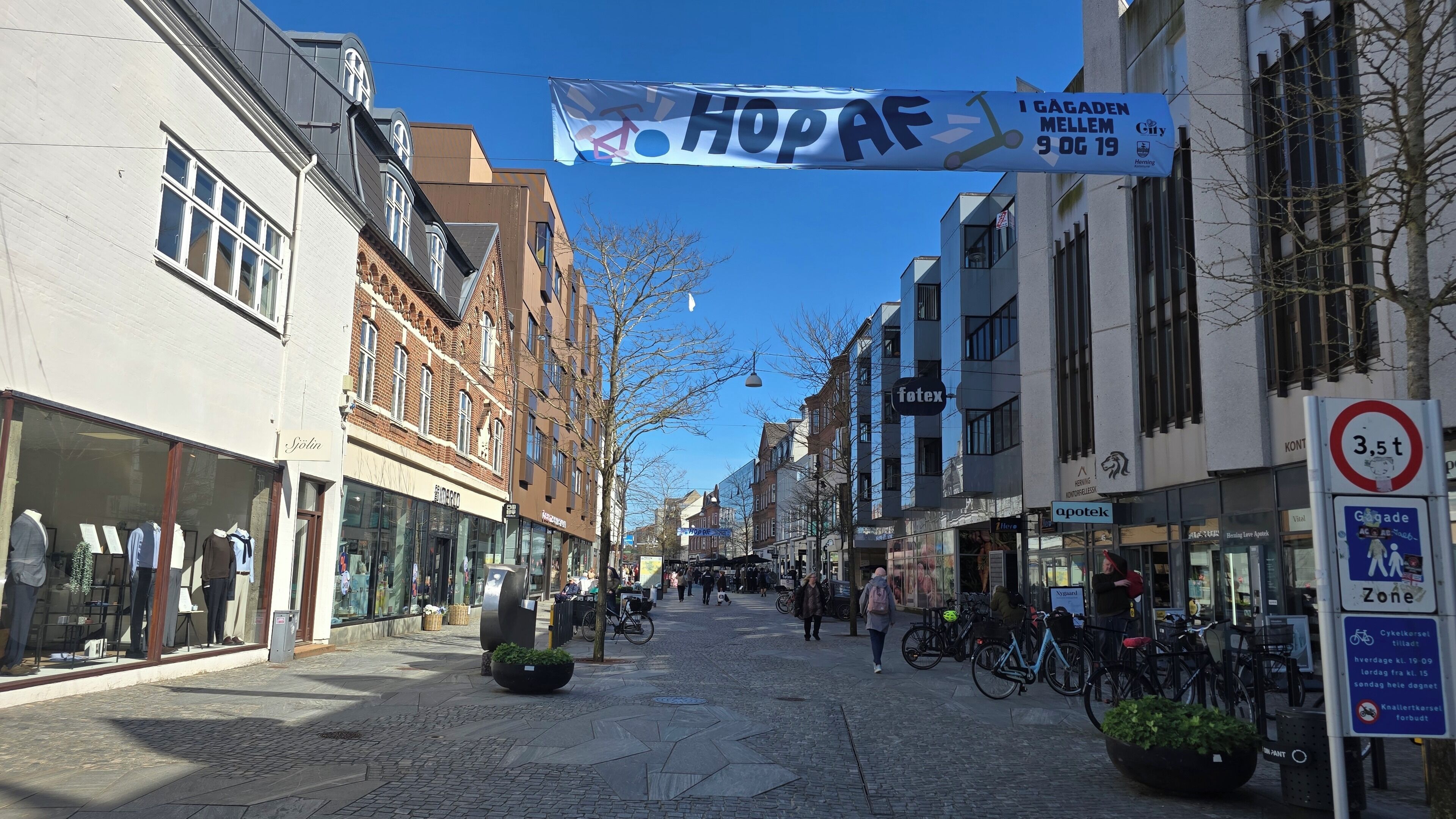 Eine gepflasterte Straße mit Geschäften und Gebäuden, ein Banner mit dem Text 'HOPAF' und ein Schild, das eine zulässige Gesamtgewicht von 3,5 Tonnen für Fahrzeuge anzeigt.