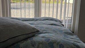 Una cama con ropa de cama estampada, una almohada y vista a través de una ventana con persianas verticales.