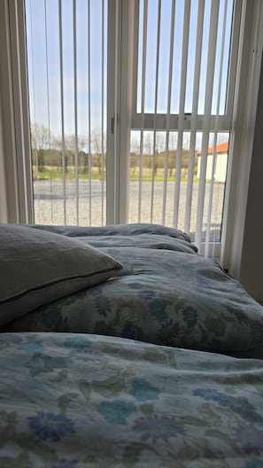 A bed with floral bedding, a pillow, and a view through a window with vertical blinds. - Sov Godt i Arnborg Bed & Breakfast (Herning)