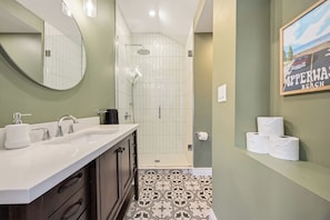 A bathroom with a white sink, mirror, and shower. There are three rolls of toilet paper on a shelf.