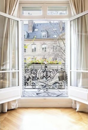 Ein Zimmer mit großem Fenster, weißen Vorhängen, einem Balkon mit Metallgeländer und Blick auf Gebäude und Bäume.
