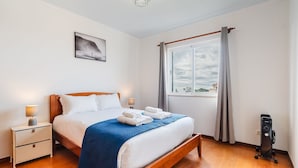 Une chambre à coucher bien rangée, avec un lit, des tables de chevet, une fenêtre avec des rideaux, un tableau accroché au mur et un luminaire au plafond.